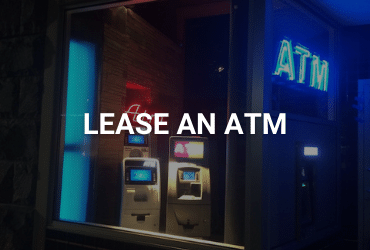 The words "Lease An ATM" are across the image in white. Night time, the photo is of the inside of an ATM store from the sidewalk. There are two ATMs lit up inside, and a bright blue neon "ATM" sign hangs in the right corner of the room. Opposite the room, is a smaller red neon "ATM" sign.