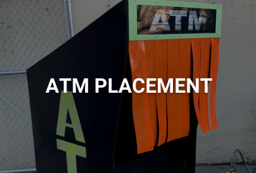 A customized outdoor ATM kiosk. the body is a reflective black and "ATM" is painted in neon green vertically along the side and horizontally along the front. There are neon orange tassels hanging for user privacy and weather protection.