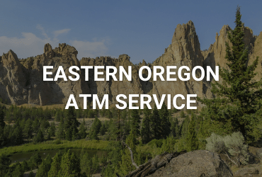 The words "Eastern Oregon ATM Service" are across the image in white. A sunny day in the rock canyons of Eastern Oregon. Large rock formations line the image, in the foreground are spiny pine trees.